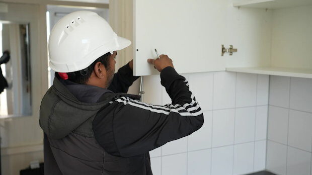 Trade worker in a white hard hat fastening a cabinet door in a kitchen.